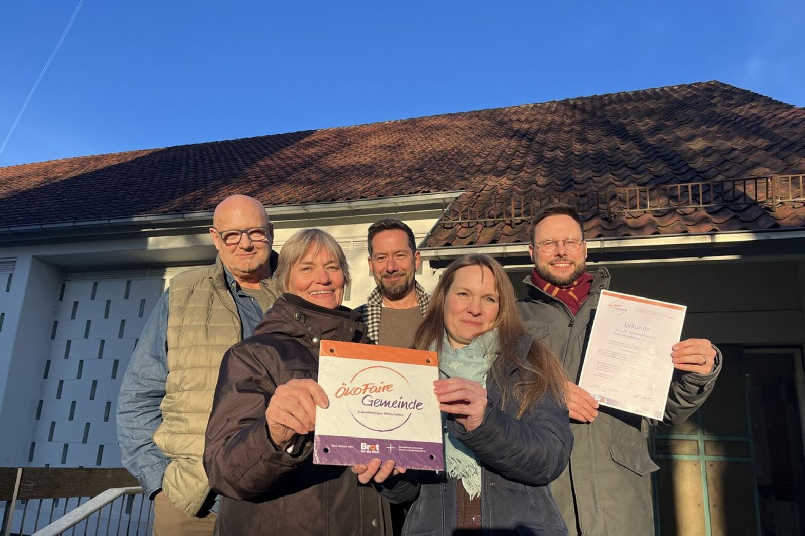 Drei Männer und zwei Frauen stehen vor einer Kirche und halten ein Schild mit der Aufschrift "Ökofaire Gemeinde"