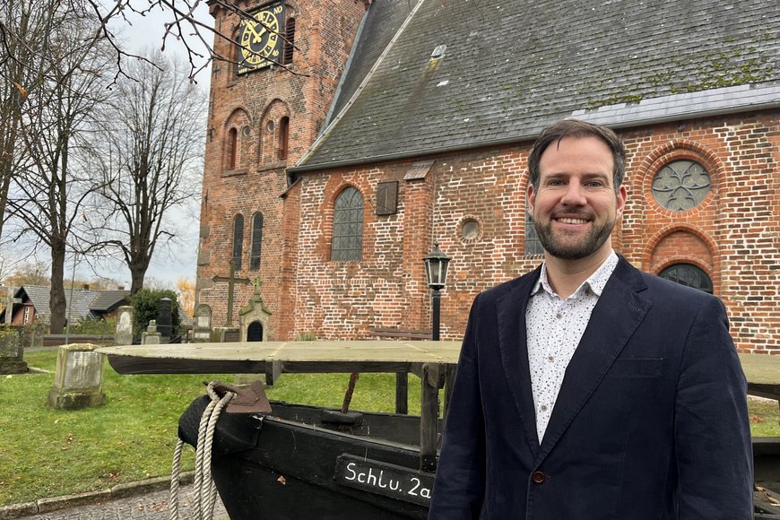 Ein Mann mit dunklem Sakko steht vor einer Backsteinkirche und lächelt in die Kamera. - Copyright: Annkathrin Bornholdt