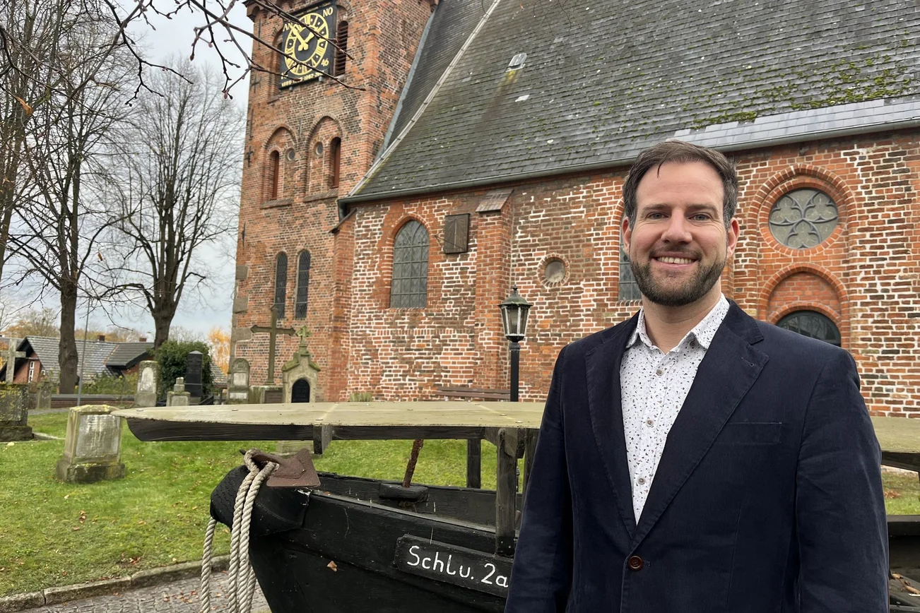 Ein Mann mit dunklem Sakko steht vor einer Backsteinkirche und lächelt in die Kamera.