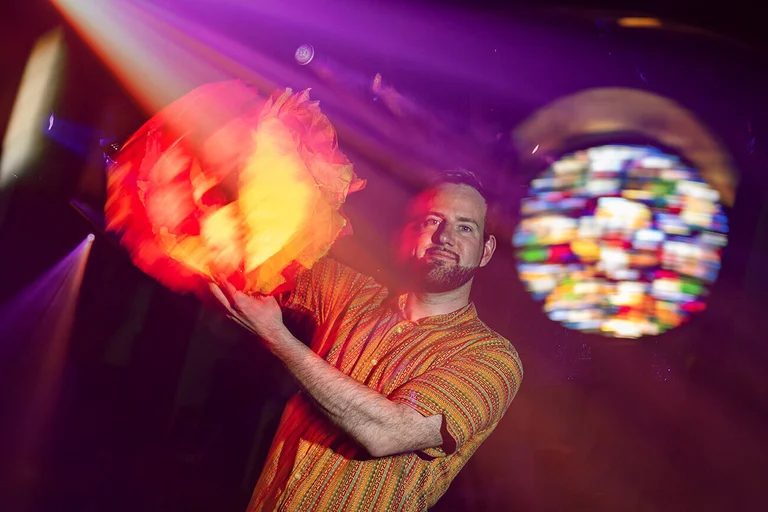 Ein Mann schaut in die Kamera. Im Hintergrund sind bunte Lichteffekte zu sehen.